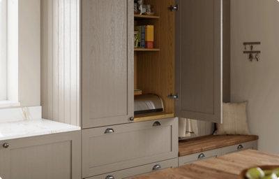 A wall-mounted shaker style kitchen cabinet in a warm grey colour is partially open, revealing glasses and boxed food. The inside of the cupboard is a warm, stained wood.