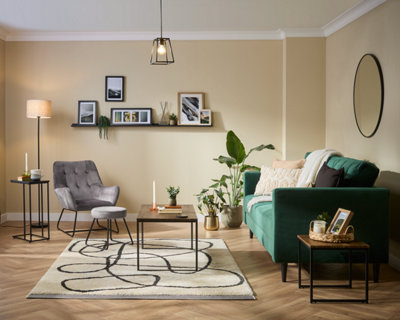 A modern living room with a green sofa, grey armchair with ottoman, minimalist coffee tables, houseplants, a white rug with black lines, framed art on a shelf, and neutral beige walls.