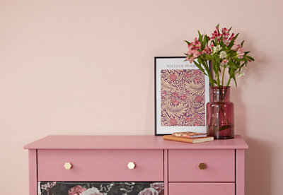 A pink cabinet with a picture, a pink glass vase with flowers, and books on top of it, standing against a pink wall.
