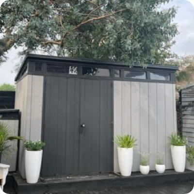 A sleek, modern garden shed with tall gray double doors and horizontal window panels near the roof is surrounded by tall white planters filled with green foliage. The shed sits on a dark patio beneath a leafy tree. This photo is user-generated content shared from a real garden on Instagram with the handle @hausonthebeach