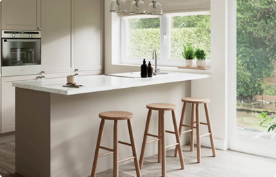 Three wooden stools sit next to a kitchen island with a white and grey marble-effect countertop. The countertop shines in the light from a nearby window. Behind, a galley kitchen features shaker-style cabinets in a warm grey shade.