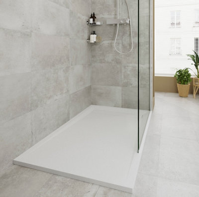 White shower tray installed in a corner between two tiled grey walls. Shower tray is rectangular with a glass shower screen along the longest side, with the rest open to create a walk-in shower