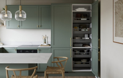 Reed green shaker kitchen with brass-effect handles, white marble-effect worktops and a matching island. Tall storage cabinet shown open to reveal internal pull-out shelving and organised pantry storage, complemented by decorative glass pendant lighting.