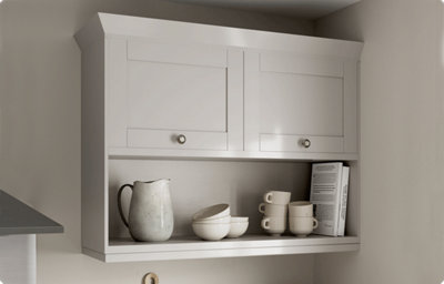 A light grey kitchen cabinet is attached to the wall. The bottom has an open shelf with books, mugs and bowls for decoration. The top of the cabinet features two shaker-style cupboards with knob handles.