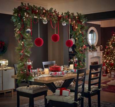 Wooden dining table with a tabletop decoration pole wrapped in a garland, featuring red ribbon, pinecones, foliage and three large red baubles hanging from the centre.