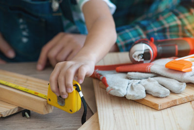 A person measuring a long piece of wood with a tape measure, there is a drill and a pair of gloves in the background.