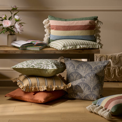 Collection of cushions in different colours and patterns stacked beneath a wooden table, including striped, botanical and textured designs in soft green, rust and neutral tones.