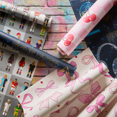 Selection of wallpaper rolls laid out on a painted wooden surface, featuring playful patterns including bows, cherries and graphic figures in pink, navy and multi-coloured designs.