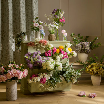 Styled interior with artificial flowers arranged on and around a green cabinet, including vases filled with mixed blooms and greenery creating a full, layered display.