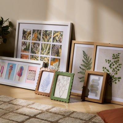 Collection of picture frames in different sizes and finishes, including wood and scalloped designs, displayed on a wooden floor with botanical and graphic prints.