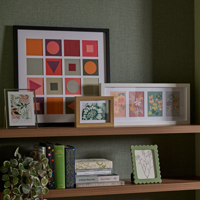 Shelving unit styled with picture frames in various sizes and finishes, displaying floral and abstract prints alongside books and trailing greenery.