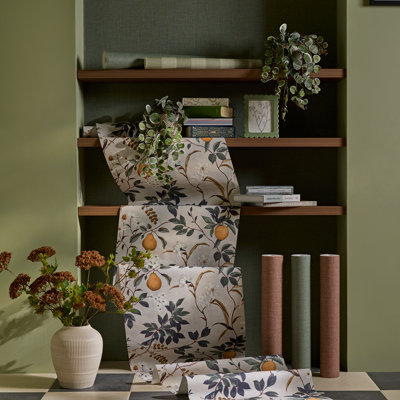 Shelving unit styled with floral wallpaper, stacked books and trailing greenery, alongside rolled wallpapers and a vase of dried flowers against a muted green wall.