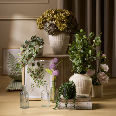 Arrangement of artificial plants and flowers in neutral ceramic vases, including trailing greenery and soft blooms, styled on stacked boxes with glass bottles on a wooden surface.