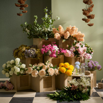 Colourful display of artificial flowers arranged in wooden crates, featuring mixed blooms in pink, peach, yellow and white tones against a green backdrop.