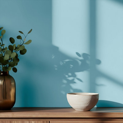 Interior detail with GoodHome Nice painted wall in a soft blue tone, featuring a wooden surface styled with a brown glass vase holding green foliage and a ceramic bowl, with natural light casting shadows across the wall.