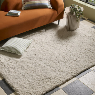 Soft cream shaggy rug styled in a living space with an orange sofa, striped cushion and side table, adding texture and warmth to the room.