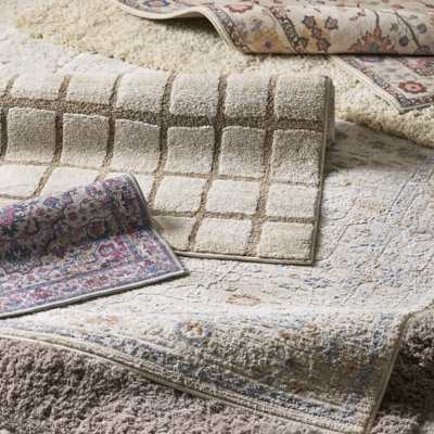 Close-up of layered rugs in neutral tones, featuring textured and patterned designs including grid and floral motifs in cream, beige and soft blue.