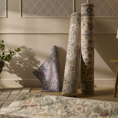 Selection of rolled rugs in a styled room, showing floral and patterned designs in soft neutral and pastel tones against a light wall backdrop.
