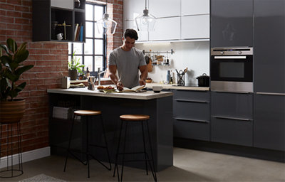 A man is reading from a book in a dark grey U-shaped kitchen. 