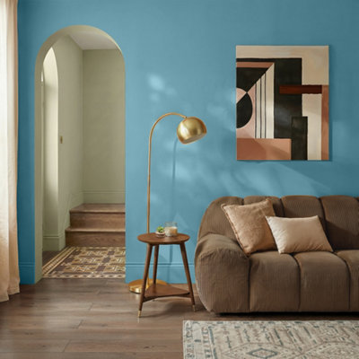 Living room with GoodHome Nice painted walls in a soft blue tone, featuring a brown upholstered sofa with cushions, a brass floor lamp, wooden side table with candle, and abstract wall art, with an arched doorway leading to a hallway with patterned flooring.