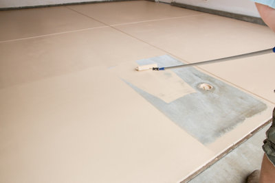 Person standing outside the garage using a roller on an extendable pole to apply paint to a garage floor.