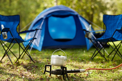 A gas powered camping stove in the middle of a camp surrounded by camping chairs and a tent in the background.
