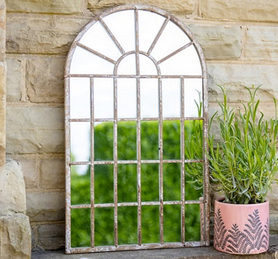 An ornate window-style arch mirror leant against an exterior brick wall.
