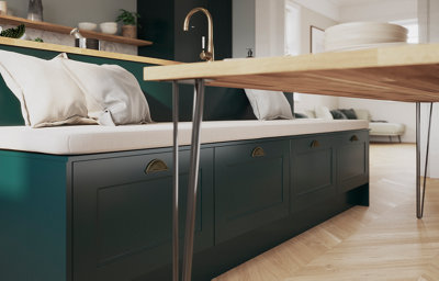 The kitchen island has bench seating with a soft, cream cushion running the length of the bench and scatter cushions are placed on top. Below, the bench has four shaker style drawers in dark green and a matt finish with gold handles.