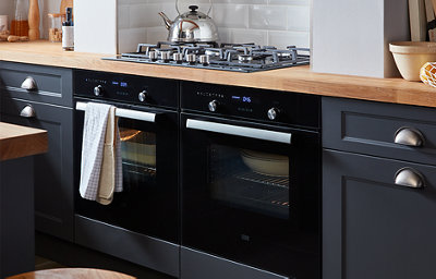 Dark grey shaker style kitchen cabinets flank two integrated ovens. The cabinets have silver coloured handles and the stained wood worktop includes a gas hob.