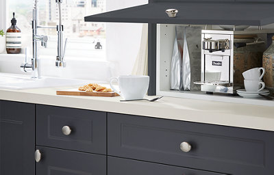 Shaker style kitchen cabinets in dark grey with a matt finish are finished with silver coloured door knobs. On the white worktop sits a fixed cabinet with a top-hinge opening. The door is lifted to reveal a coffee machine.