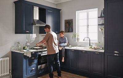 A man is cooking on the hob in an L-shaped kitchen with dark blue, matt finish, shaker style cabinets. A child sits on top of the white marbled kitchen worktop.