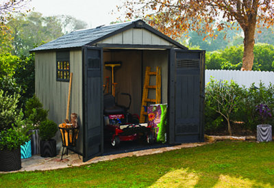 A large plastic shed installed in a garden, surrounded by a bright green lawn and various plants.