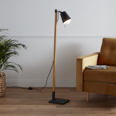 Floor lamp with black floor plate, wood vertical support and black lamp shade. Lamp is placed on wood flooring in front of white wall.