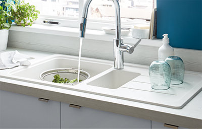 Modern white inset sink with chrome mixer tap and drainer, set in a light wood-effect worktop. Features fresh vegetables being rinsed and glassware drying on the side.
