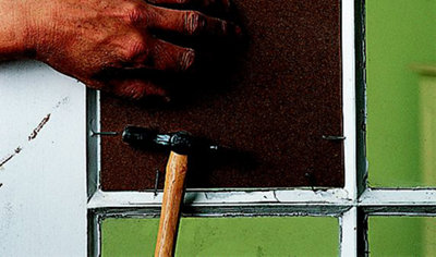 Using a hammer to drive a nail into a piece of timber, securing it in place over a hole where a pane of glass has been broken.