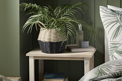 Small plant in a rattan plant pot sat on a bedside table. Bedside table is next to a bed with nature inspired sheets.