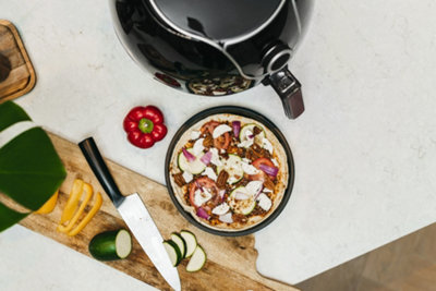 An air fryer placed on a kitchen worktop next to a chopping board, fresh vegetables, and a homemade pizza.