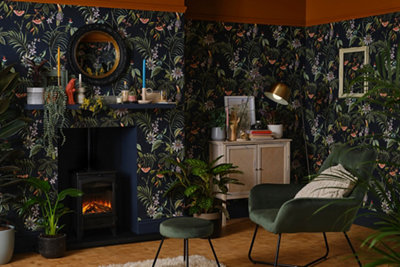A living room decorated with dark blue botanical print wallpaper, a green armchair and footstool, and a variety of houseplants.
