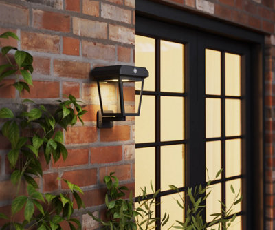 A small black outdoor wall light with a light at the top of the fixture, projecting down into the square clear frame below.