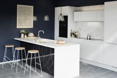 A kitchen with dark blue walls, white cabinets and white worktops, with a large white breakfast bar with 3 bar stools.