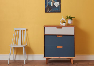 A cabinet with grey and white drawers and a white chair against the background of a yellow wall with a picture hanging on it.