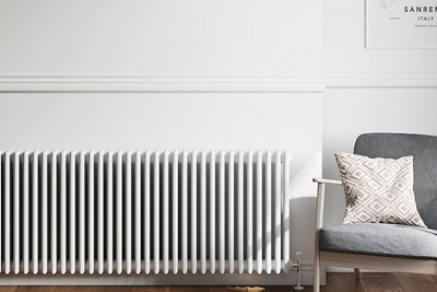 A white radiator against a white wall, next to which stands a grey armchair with a light grey and white cushion.