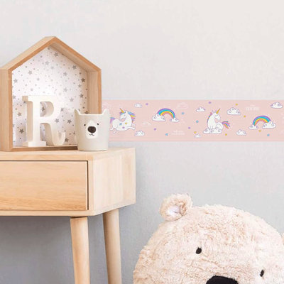 A child's bedroom decorated with a large teddy bear, a wooden desk and neutral walls featuring a pink, unicorn themed wallpaper border.