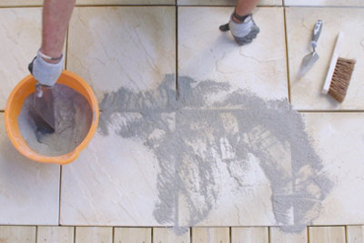 Person wearing gloves scooping up a mixture of sand and cement from a bucket with a trowel. The mixture has already been spread into the gap between two paving slabs.