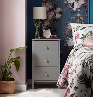 A grey wooden bedside table with legs besides a bed with a bold floral duvet cover.