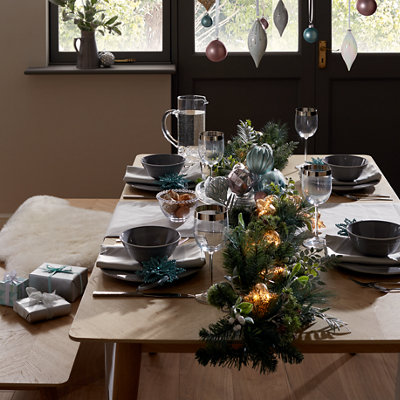 Wooden dining table decorated for Christmas with a festive garland of baubles, lights and foliage down the centre, set with crockery, napkins and glasses.
