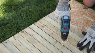 Person wearing gloves using a drill to drill pilot holes in pieces of timber which are lined up. Line has been drawn across the pieces of timber showing where pilot holes need to be drilled.
