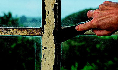 Using a scraper to remove flaking paint from a rusted metal window frame.