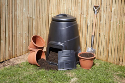 A compost bin positioned in the corner of a garden, next to flower pots and a shovel.