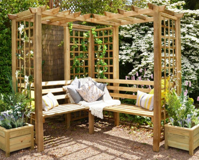 A wooden corner pergola with a built-in bench, decorated with outdoor cushions and blankets for a cosy retreat.